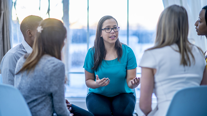 A multi-ethnic group of adults are attending a mental health support group. A woman is sharing her problems while they sit in a circle.